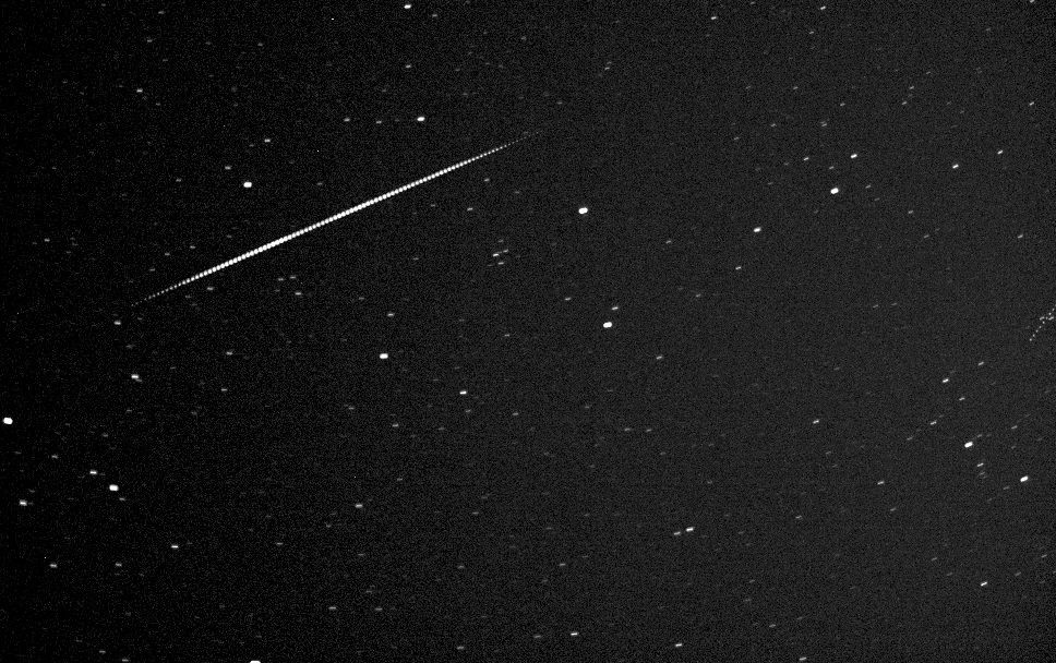 A satellite crosses a field of stars as a dashed trail of about a hundred short strokes, running from the left edge up towards the middle of the frame. The stars around it stay points.